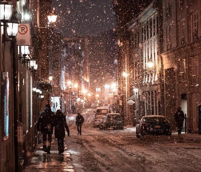 Two people with backs to the camera walk down a city sidewalk at night in a snowstorm.